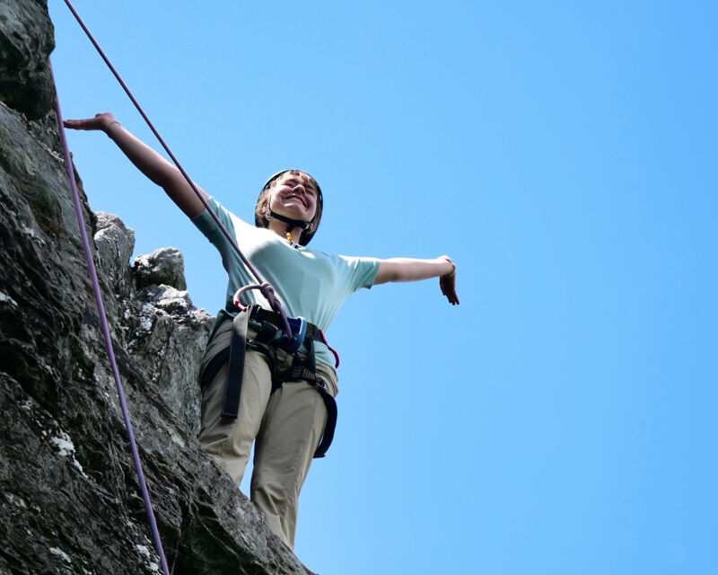A woman is rock climbing, wearing a helmet and harness. She's on a rocky cliff face with a rope extending upwards. Her arms are outstretched, and she appears to be smiling or laughing, possibly celebrating a successful climb. The sky is a clear, bright blue in the background.